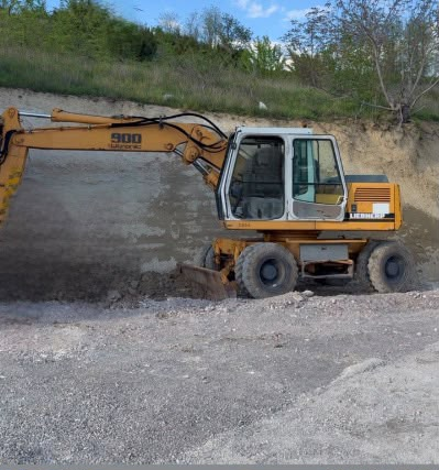Ekskavator i verdhë i ndalur mbi një sipërfaqe me zhavorr para një pjerrësie toke