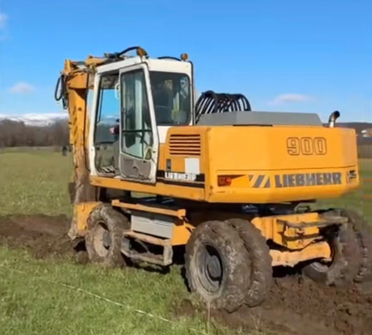 Ekskavator i verdhë që hap një kanal në një livadh me male në largësi